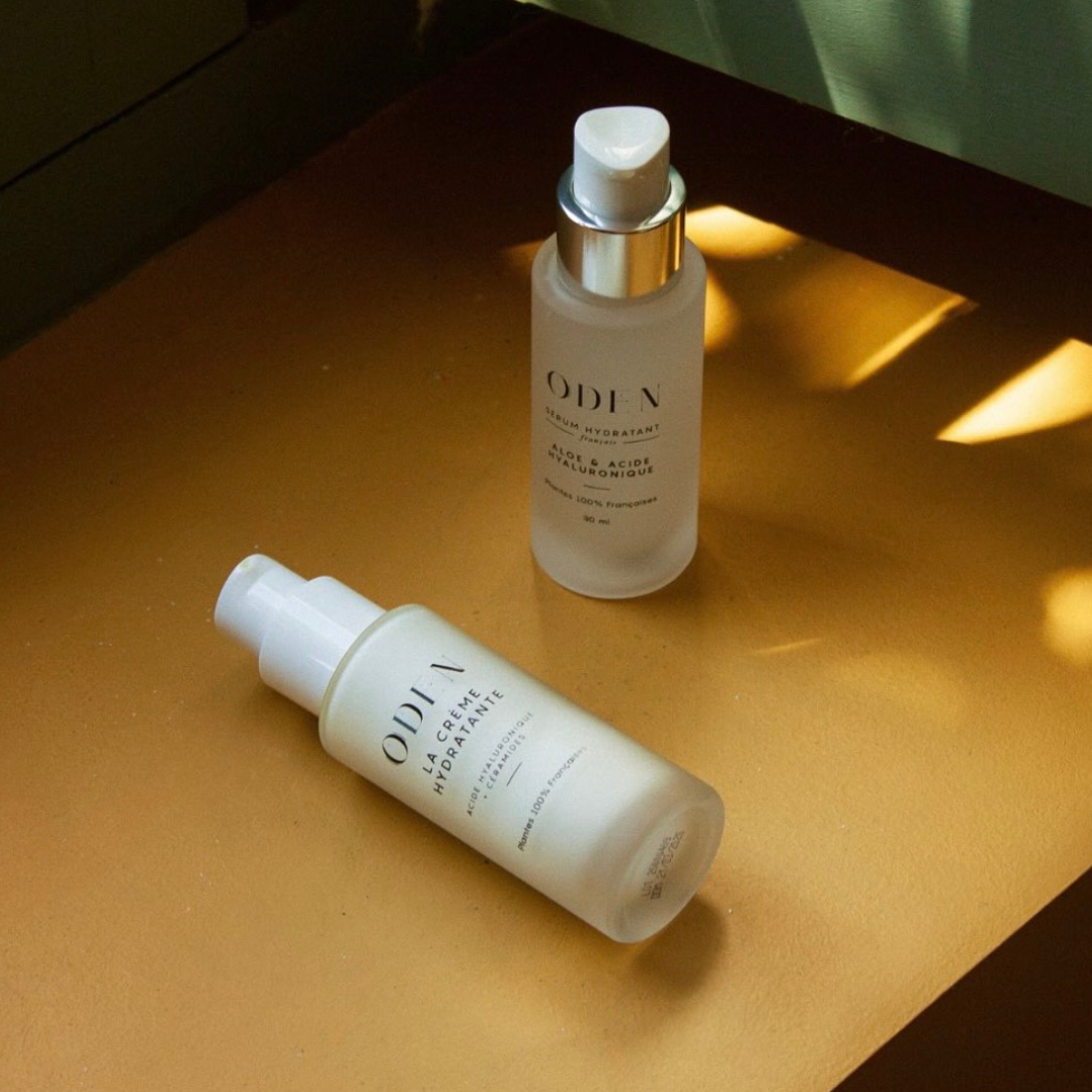 Two ODEN skincare bottles, one with a white pump bottle and one with a frosted glass dropper bottle and a gold collar, resting on a warm brown surface with dramatic golden sunlight and shadows.