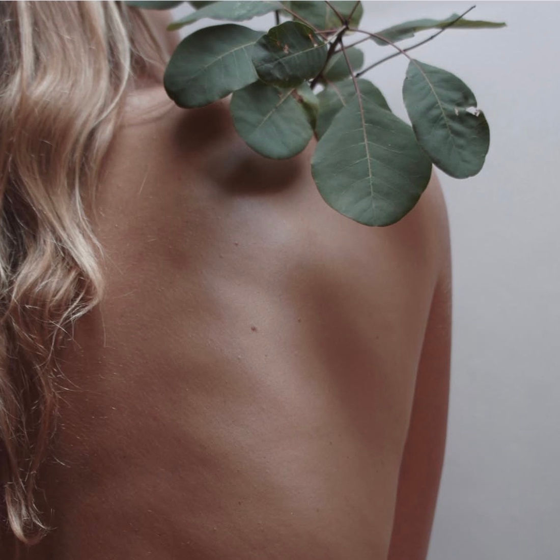 A close-up lifestyle shot of a woman's bare back and shoulder, with blonde hair on the left and green eucalyptus leaves resting on her upper back against a soft gray background.