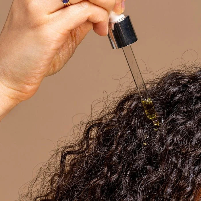 Hand applying a dropper of oil to curly hair against a beige background